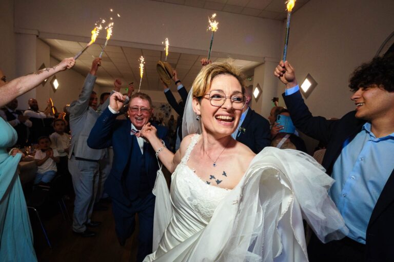 Mariés entrant dans la salle de réception sous une haie d'étincelles, rieurs et complices, entourés de leurs invité·es en liesse – photo prise lors d’un mariage festif en intérieur.