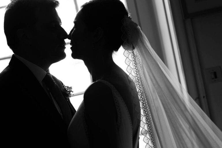 Couple de marié·es en contre-jour, la mariée porte un voile en dentelle et le couple s’apprête à s’embrasser.