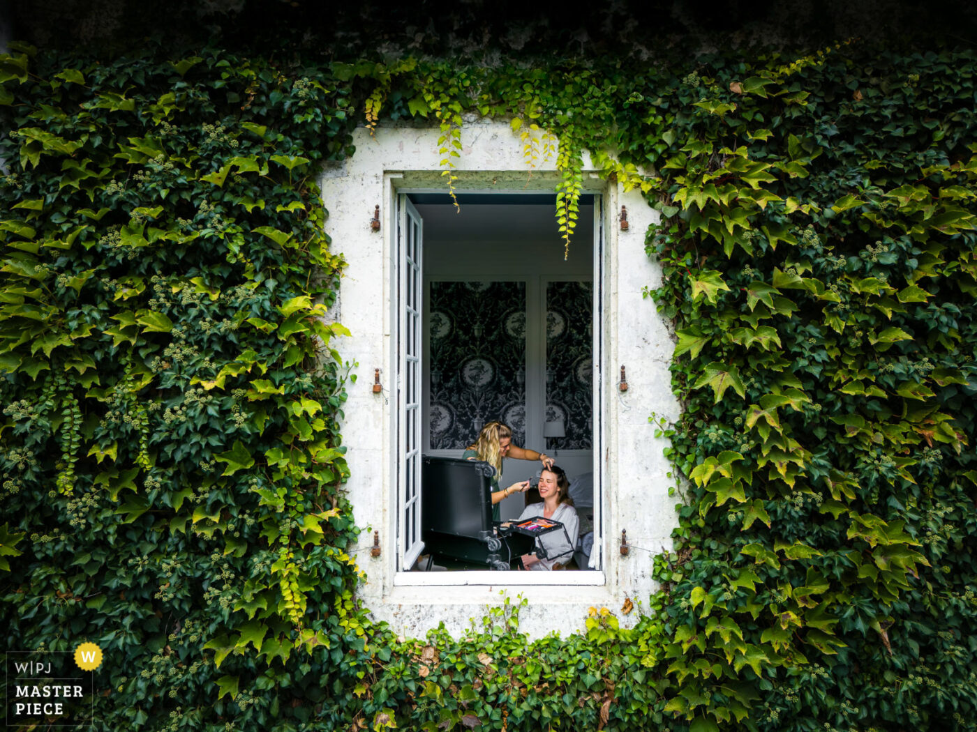 Mariée en préparation vue à travers une fenêtre entourée de lierre avec maquillage en cours